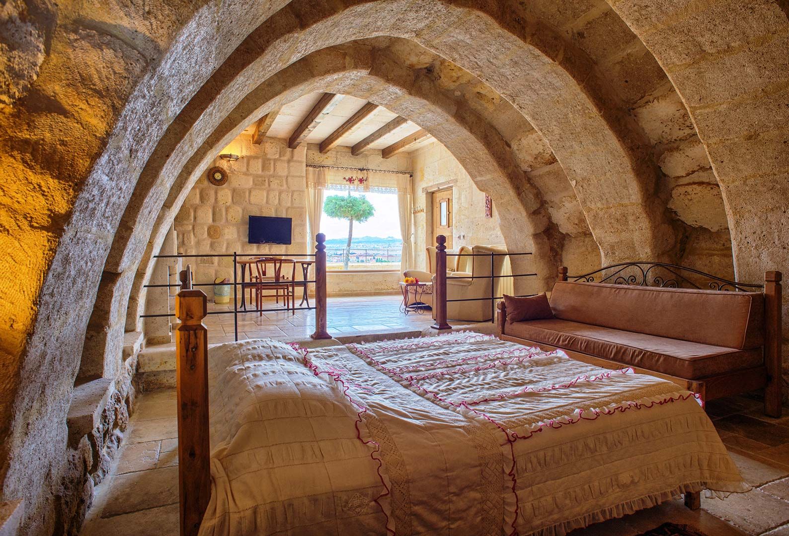 A bedroom with a stone archway, a bed with a quilt, a wooden bench, and a view of the outdoors.