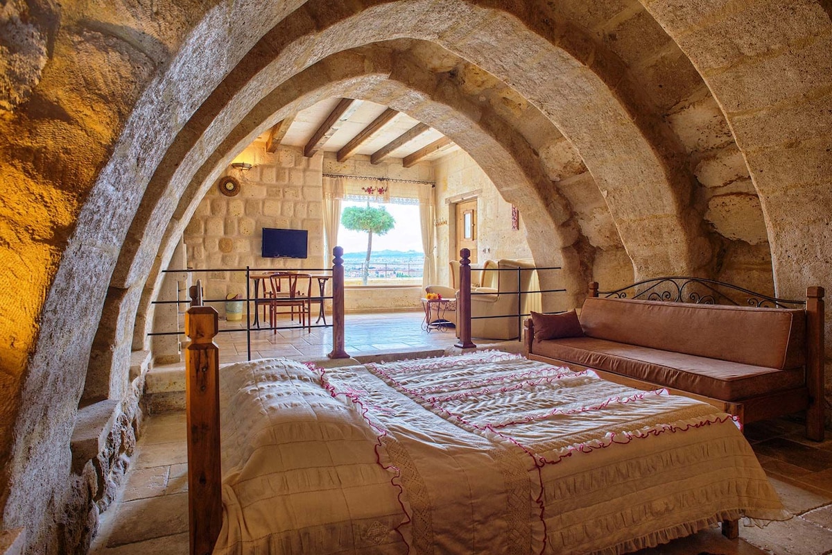 Un dormitorio con un arco de piedra, una cama con colcha estampada, un banco de madera y vistas al exterior.