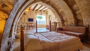 Quarto com arco de pedra, cama com colcha, banco de madeira e vista para o exterior.