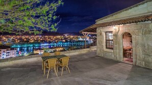 Um terraço com vista para uma paisagem urbana iluminada à noite, um edifício de pedra com uma janela e uma mesa com cadeiras.