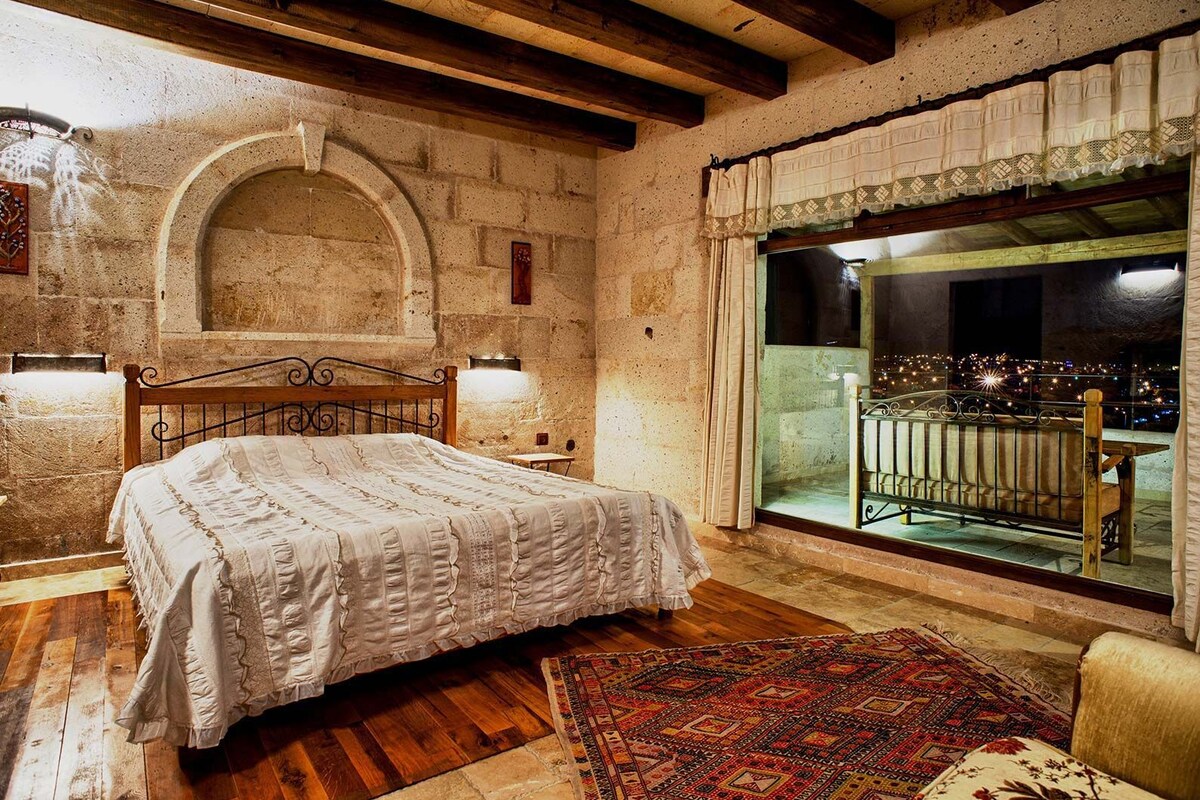 A bedroom with a wooden bed, a large window with a balcony, stone walls, and a rustic wooden ceiling.