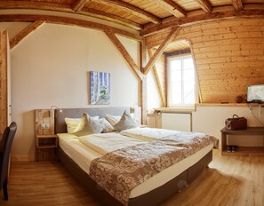 Un dormitorio con techo de madera, una cama con colcha estampada, una mesita de noche con lámpara, un cuadro en la pared y un televisor sobre una estantería.