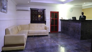A reception area with a beige sofa, a red door, a reception desk, and a clock on the wall. - Nelseno Hotel (Abuja)