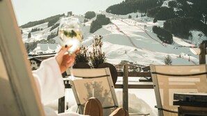 Una persona sosteniendo una copa de vino blanco, sentada en una mesa con vista a una pista de esquí nevada y edificios al fondo.