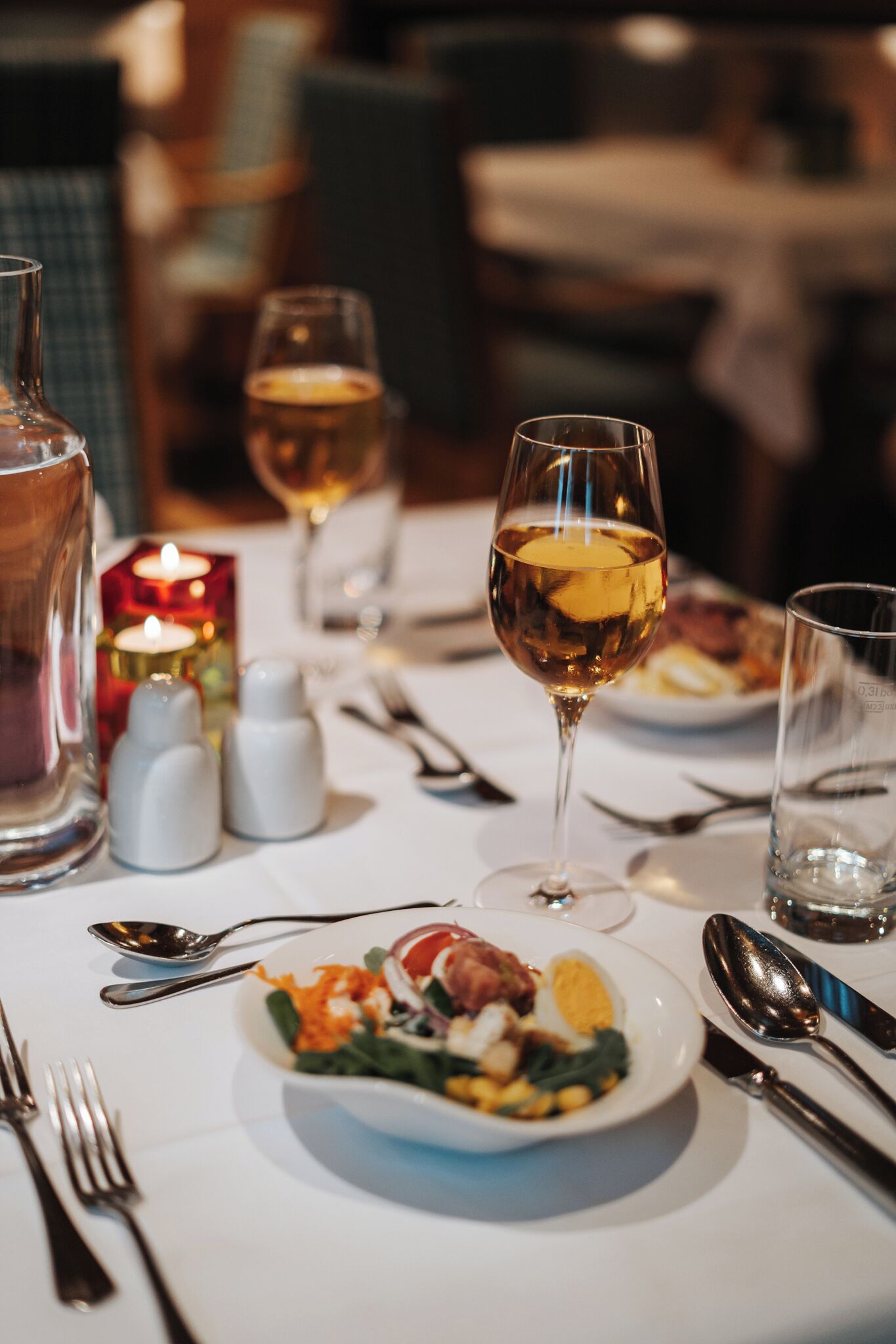 Una mesa de comedor dispuesta con una copa de vino blanco, saleros y pimenteros, una vela y un plato de comida.