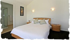 Une chambre avec un grand lit, deux tables de chevet, une commode, un tableau encadré au mur et une porte donnant sur une autre pièce.