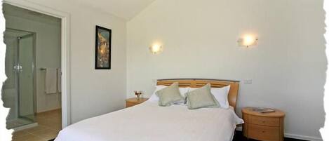 A bedroom with a large bed, two bedside tables, a nightstand, a framed picture on the wall, and a door leading to another room.