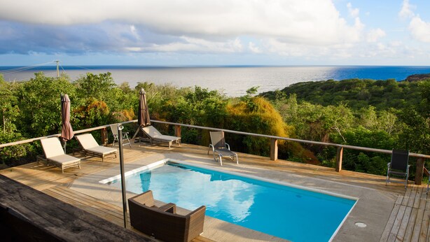 A swimming pool with lounge chairs and umbrellas on a wooden deck overlooking a body of water and greenery. - The Cottage, 3 Bedroom Rustic Villa (Belle Isle)