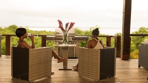 Two people seated on wicker chairs facing a grill and a table with a vase of flowers on a wooden deck with a view of the ocean. - The Cottage, 3 Bedroom Rustic Villa (Belle Isle)