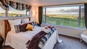 Ein Schlafzimmer mit einem großen Fenster, das einen Blick auf sanfte Hügel bietet. Das Bett ist ordentlich gemacht mit vielen Kissen und einer Decke.
