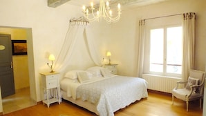 A bedroom with a bed, two bedside tables with lamps, a chair, a window with curtains, and a chandelier. - La Royante (Aubagne)