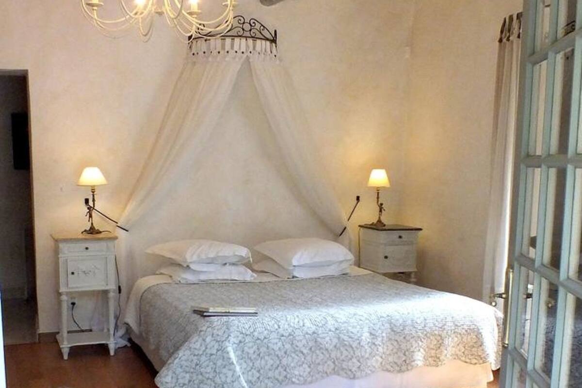 A bedroom with a canopy bed, two bedside tables with lamps, a chandelier, and a wooden floor.