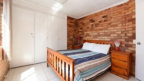 A bedroom with a wooden bed, a brick accent wall, a white wardrobe, and a bedside table with a lamp.