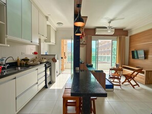 A modern kitchen with a central island, a dining area with wooden chairs, and a TV mounted on the wall.