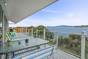 A deck with a glass table and chairs, overlooking a body of water and greenery. - Dolphin View Beach House (Primrose Sands)