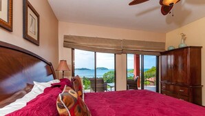 A bedroom with a large bed, a wooden headboard, a nightstand, a lamp, and a view of the sea through large windows.
