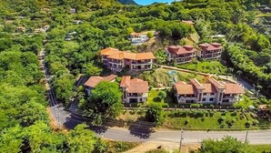 A hillside residential area with multiple houses featuring red-tiled roofs and white walls, surrounded by lush greenery and a clear road.
