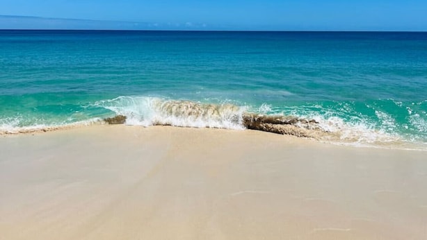 Une plage bordée d’une eau turquoise et limpide, avec un rivage sablonneux, le tout sous un ciel bleu parsemé de quelques nuages.