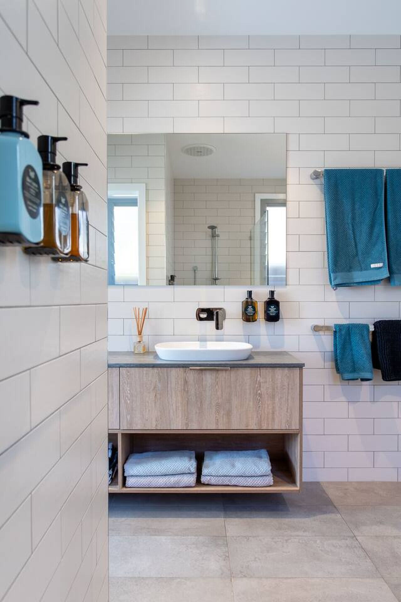 Un baño moderno con un lavabo blanco, tocador de madera y un espejo grande. Hay dos toallas colgadas en un perchero y un estante con artículos de aseo.