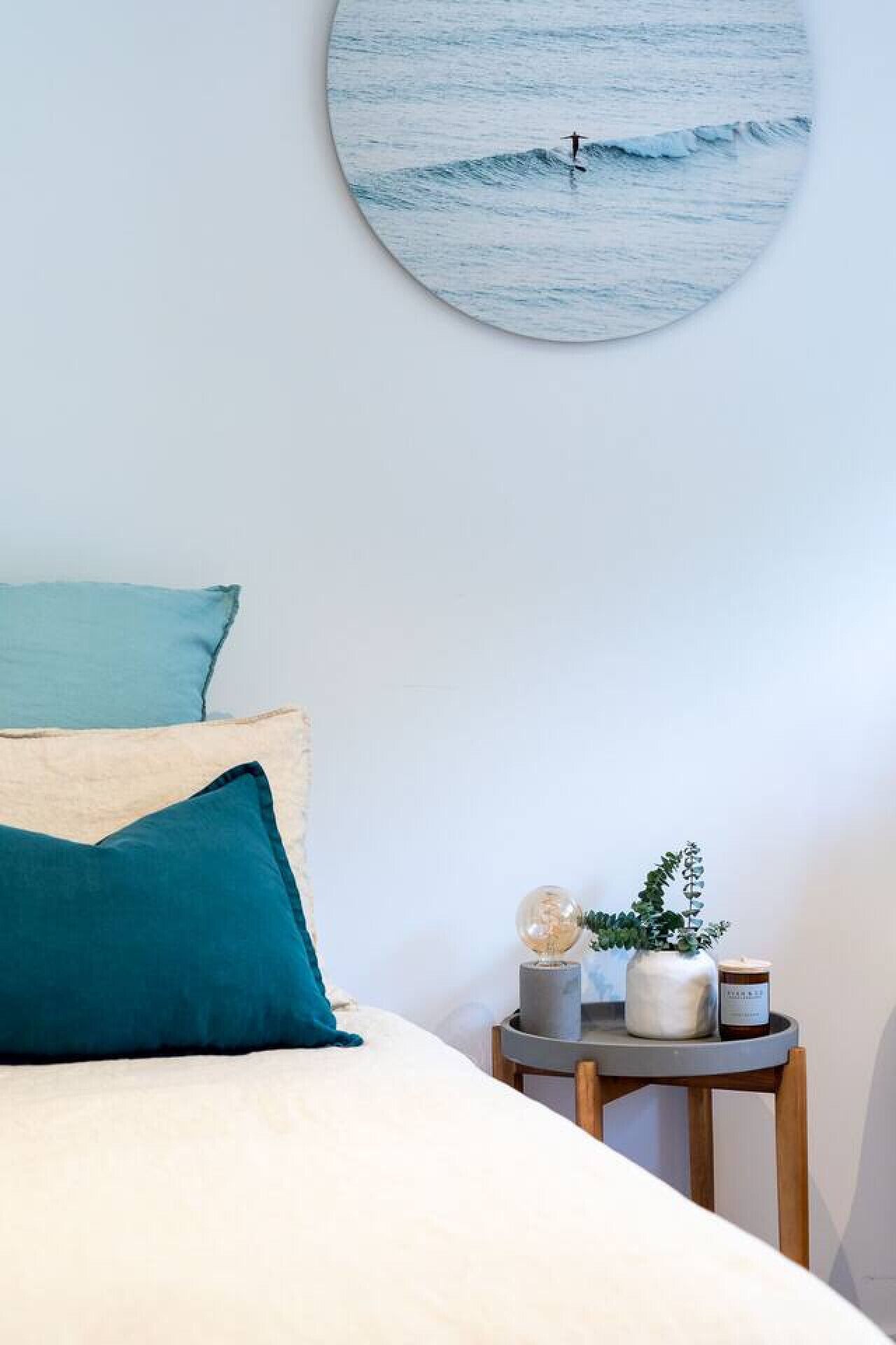 Una cama bien hecha con dos almohadas, un cuadro redondo de un surfista y una mesita de noche con una planta y una vela.
