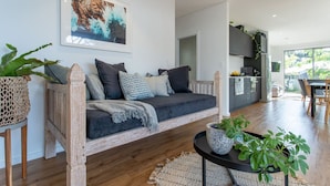 Un salon moderne avec un canapé gris foncé, des fauteuils à armatures en bois et une table basse agrémentée de plantes. On aperçoit en arrière-plan une cuisine avec des meubles gris et un coin repas composé d’une table et de chaises.