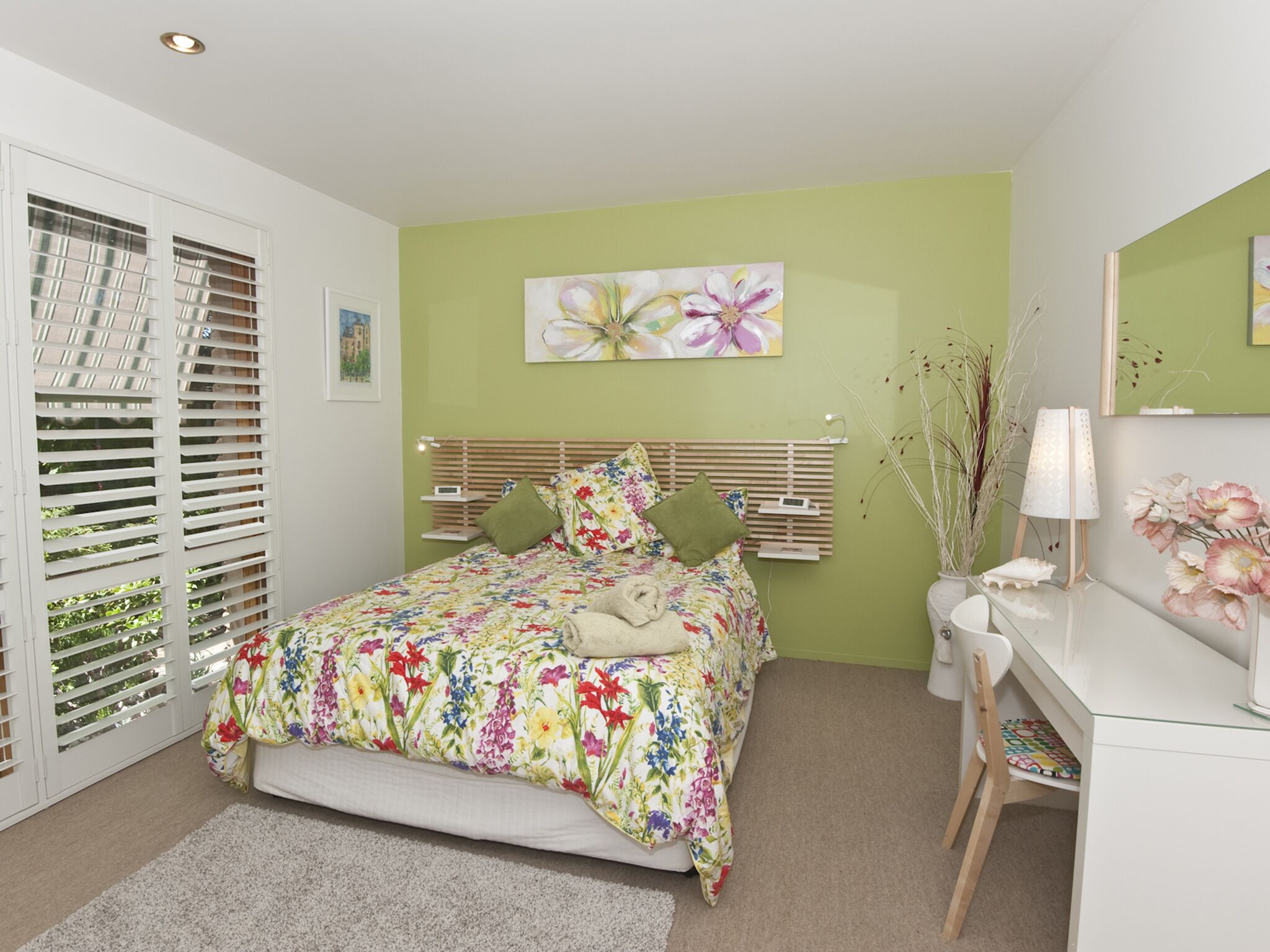 A bedroom with a bed, bedside table, lamp, and a desk with a chair.