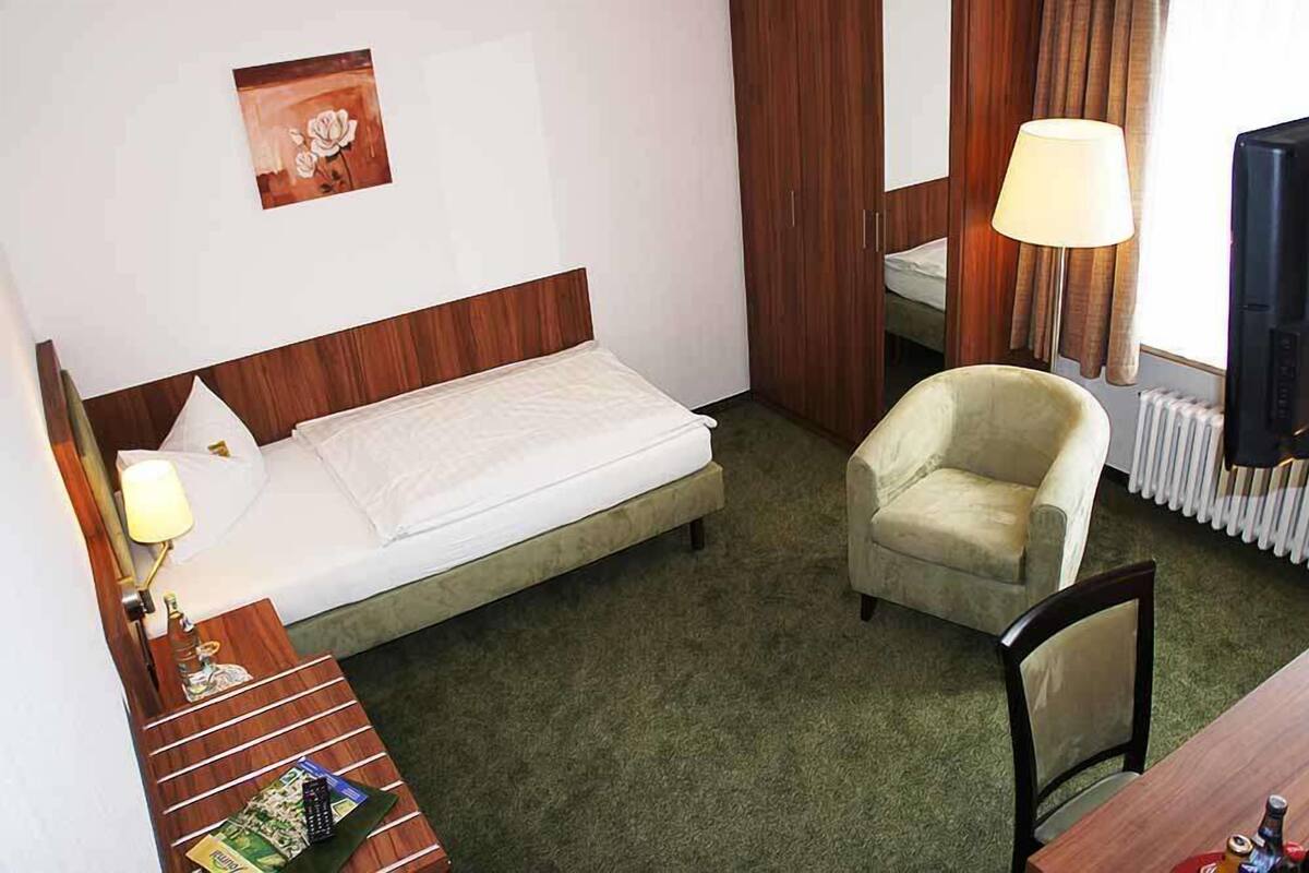 Ein Hotelzimmer mit einem Einzelbett, einem Sessel, einem Schreibtisch mit Lampe und einem Bild an der Wand.