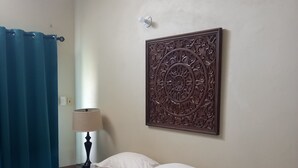 A bedroom with a bed, a nightstand, a lamp, a decorative wall piece, and a blue curtain. - Easy Walk To Store Bay, Pigeon Point, Restaurants & Entertainment (Crown Point)