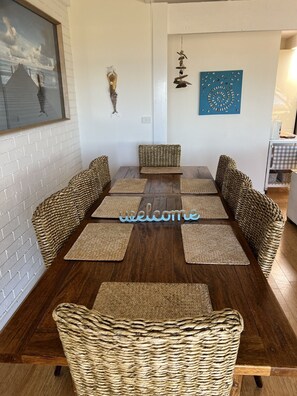 A dining area with a wooden table and woven chairs, a 'welcome' sign, and wall art. - The Esplanade Beach House (Portarlington)
