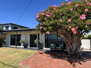 A modern house with a large tree in the front yard, a patio with chairs, and a clear blue sky. - The Esplanade Beach House (Portarlington)
