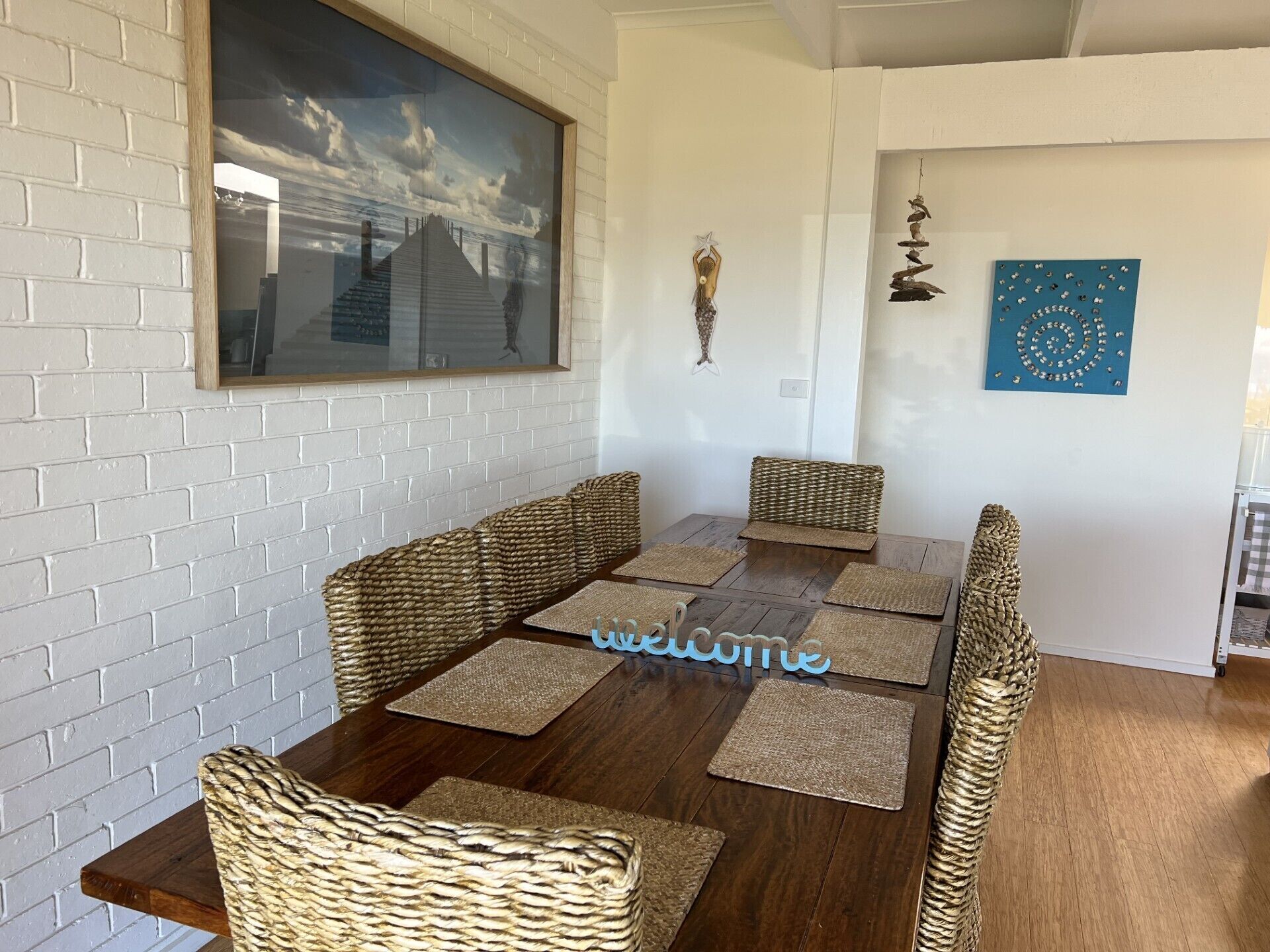 A dining area with a wooden table and woven chairs, a framed picture on the wall, and a decorative piece above the table.