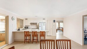 A modern kitchen with white cabinets, a microwave, and a refrigerator. There are wooden chairs around the kitchen island. - Home away from home - Close to the beach (GERRINGONG)