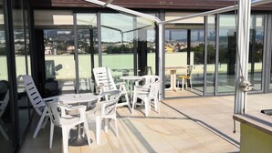 A covered outdoor seating area with chairs and tables, large windows, and a view of the cityscape. - Hotel Villa Mary (Loano)