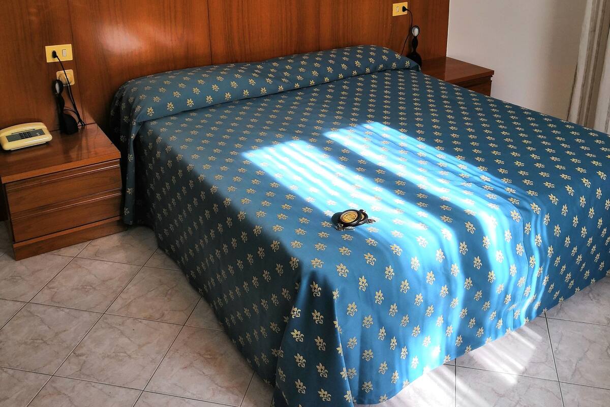 A bedroom with a wooden headboard, a bed with a blue floral bedspread, a bedside table with a telephone, and a wall-mounted light switch.
