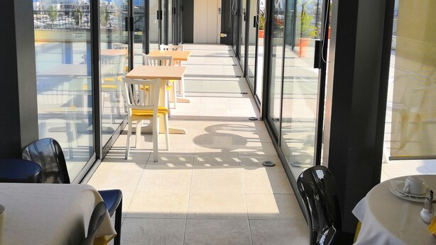 A corridor with tables and chairs, large windows, and a view of the outside. - Hotel Villa Mary (Loano)