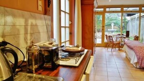 A kitchen with a stove, kettle, and coffee maker, a dining area with a table and chairs, and a view of a garden through large windows. - Melbourne Street Mews Spa Cottage (Adelaide City)