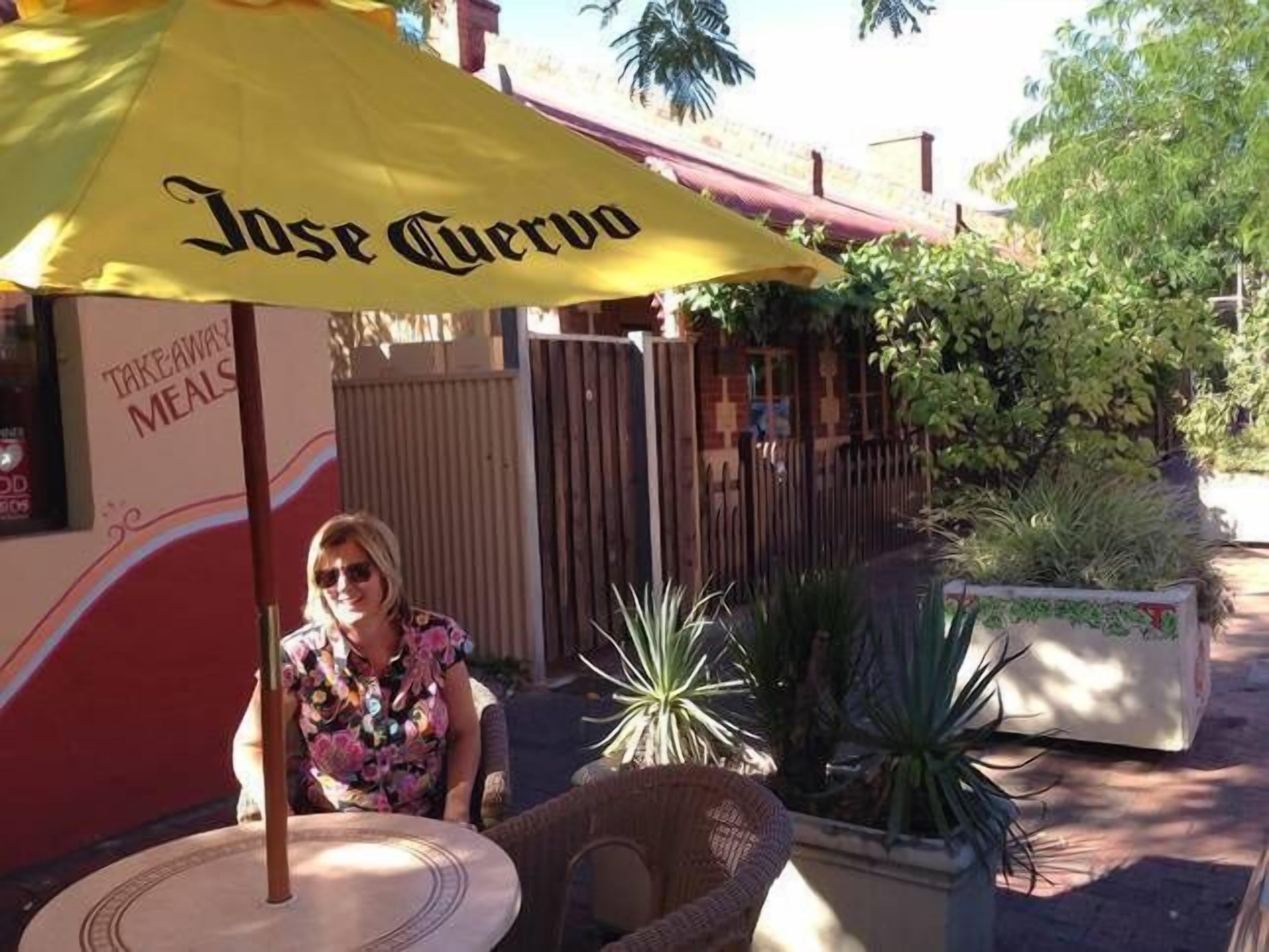 A person seated under a yellow umbrella with "Jose Cuervo" written on it, outside a restaurant with a sign for "Takeaway Meals".