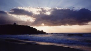 A coastal scene with a lighthouse on a hill, waves crashing onto the shore, and a cloudy sky. - STRATHMORE LODGE  UNIT 1. UPPER LEVEL. Spectacular views of Lighthouse Beach. (Port Macquarie)