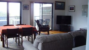 A living room with a dining table, chairs, a sofa, a TV, and a balcony with a railing. - STRATHMORE LODGE  UNIT 1. UPPER LEVEL. Spectacular views of Lighthouse Beach. (Port Macquarie)
