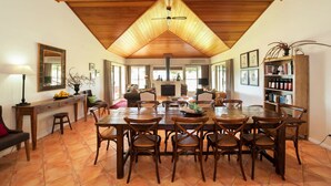A spacious dining area with a high wooden ceiling, a central table surrounded by chairs, and a kitchen area in the background.