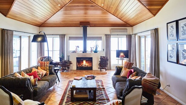 A spacious living room with a high wooden ceiling, a fireplace, and large windows.