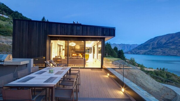A modern house with a deck overlooking a lake, featuring a dining area and a fireplace.