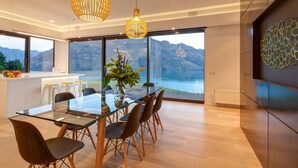 A modern dining area with a glass table, wooden chairs, and a view of mountains and a lake through large windows.