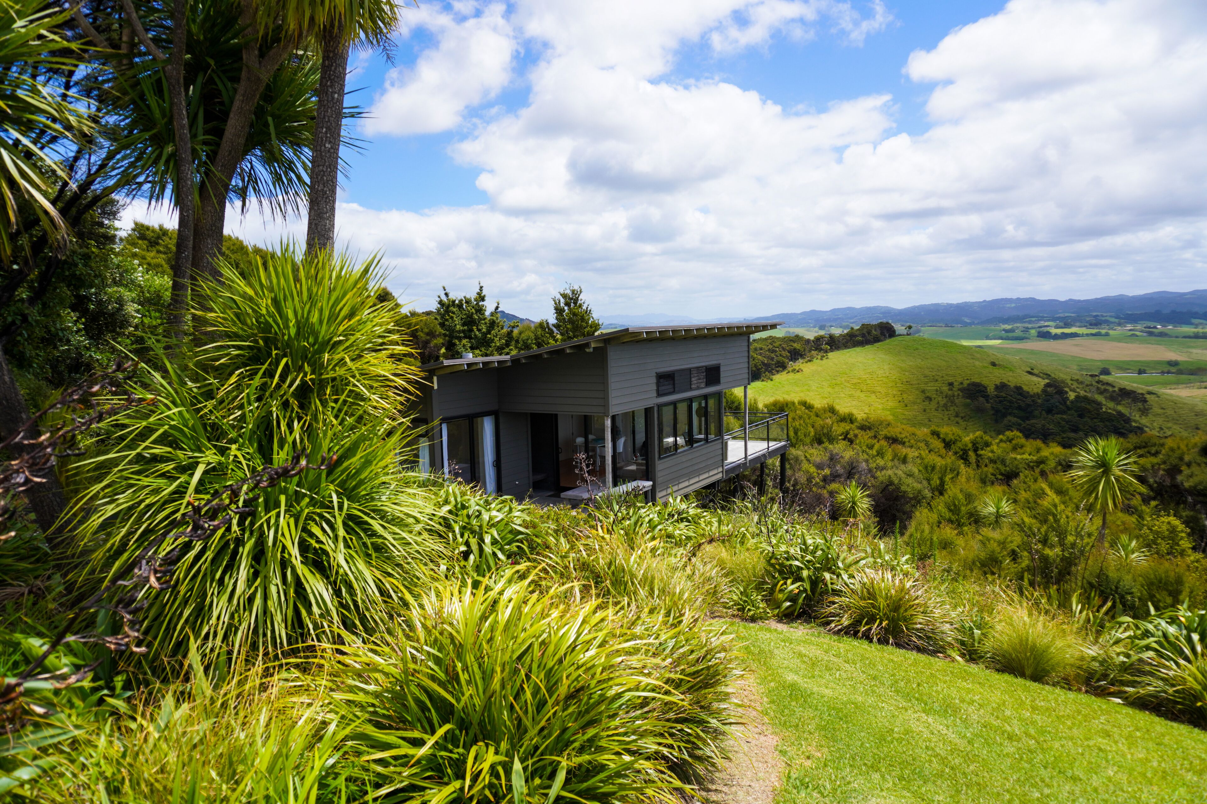 A modern house with a glass facade, surrounded by lush greenery and a hilly landscape.