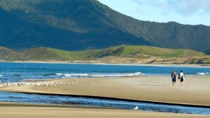 Una playa con dos personas caminando, una colina rocosa al fondo y una bandada de pájaros cerca del agua.