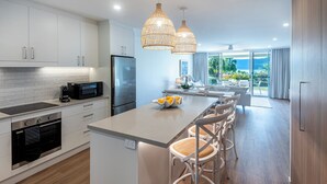 A modern kitchen with white cabinetry, a central island with bar stools, a built-in oven, and a microwave. - Lagoon 006 Hamilton Island by HamoRent (Hamilton Island)