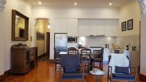 A spacious living area with wooden flooring, a dining table with chairs, a small round table with a vase, and a kitchen in the background.