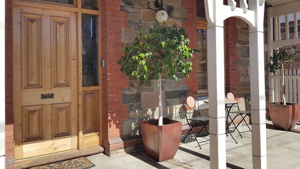 A wooden door, a stone wall, a potted tree, a white porch, and a table with chairs.