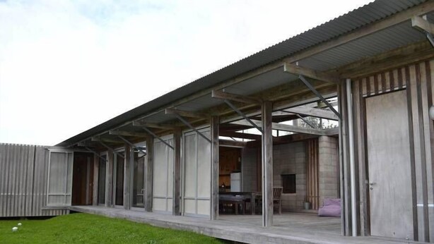 A modern, single-story building with a flat roof and large windows. It features a covered outdoor area with a table and chairs.