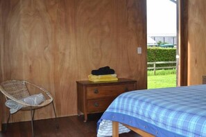 A room with a bed, a wooden dresser, a wicker chair, and a view of a green landscape. - Modern three bedroom house, located at Medlands, Great Barrier Island. (Medlands Beach)